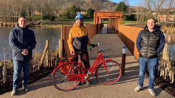 Conor Maskey and Winston Irvine from Intercomm standing either side of Sustrans Rachael Ludlow-Williams with her bike on the new greenway with footbridge at Springfield Dam on the Forth Meadow Community Greenway in the background